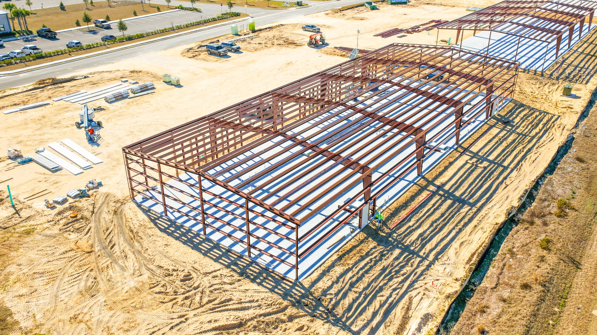 Aerial view of a construction site with a row of steel-framed structures on sandy ground, casting long shadows under a bright blue sky.