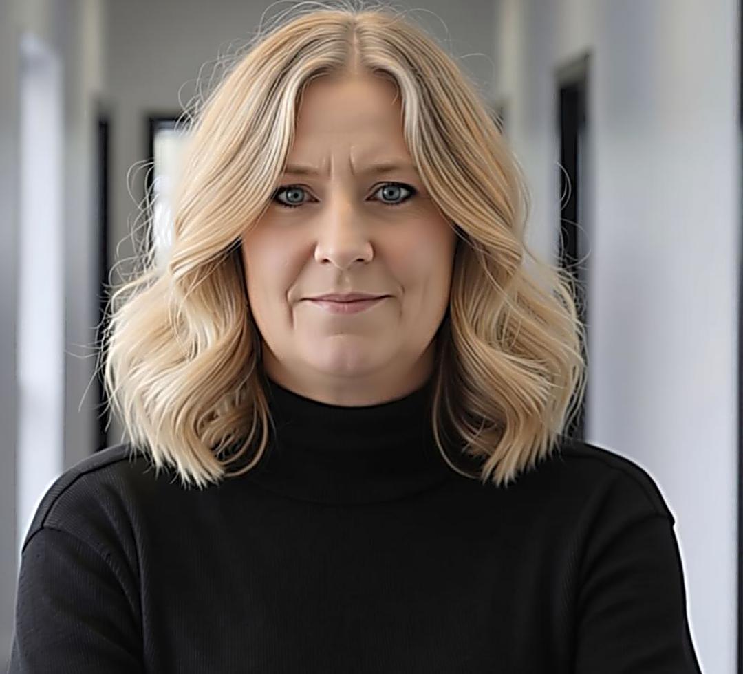 Woman with wavy blonde hair wearing a black turtleneck stands confidently in a hallway, conveying professionalism and approachability.
